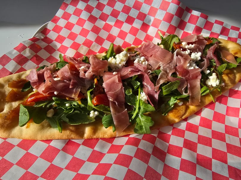 Flatbread with prosciutto, arugula, goat cheese, and sun-dried tomatoes on red and white checkered paper.