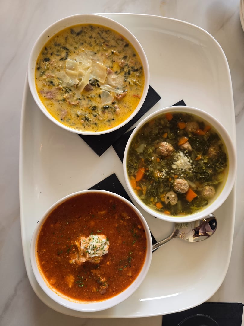 Three bowls of soup on a white platter: creamy yellow, green with meatballs, and red with a dollop of cream.