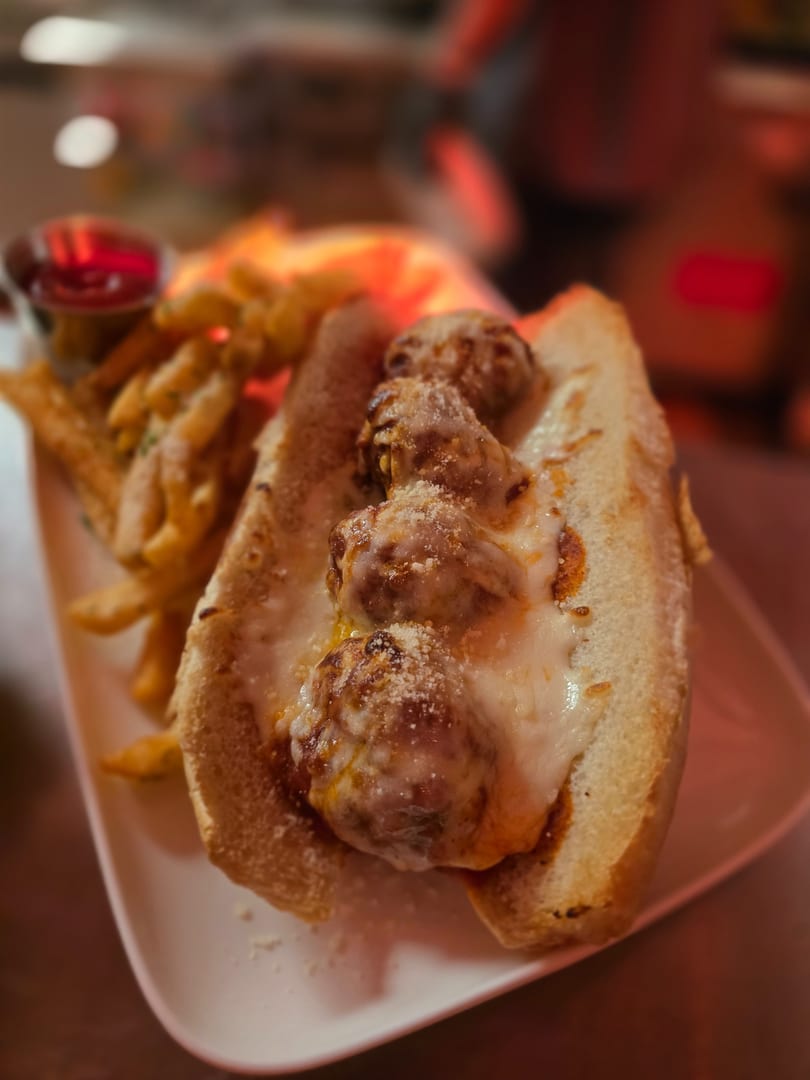 Meatball sub sandwich with melted cheese and fries on a white plate, with a red dipping sauce.