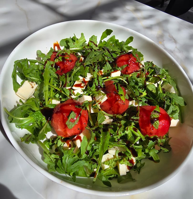 Arugula salad with watermelon, feta cheese, mint, and balsamic glaze in a white bowl.