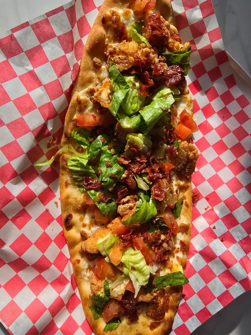 A crispy flatbread pizza topped with fried chicken, bacon, lettuce, and tomatoes on checkered paper.